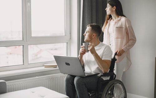 Чоловік в інвалідному візку з ноутдуком на колінах і дівчина позаду дивляться у вікно | © Pexels